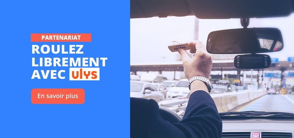Bannière de partenariat avec Ulys. Visuel pris depuis l’intérieur d’un véhicule montrant une main tenant un badge de télépéage à l’approche d’un péage autoroutier. Texte « Roulez librement avec Ulys ». Bouton d’appel à l’action « En savoir plus ».